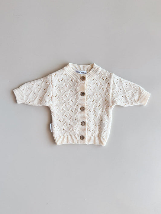 A cream-colored baby knit cardigan sweater with a diamond lattice pattern and brown buttons, laid flat on a light gray background.