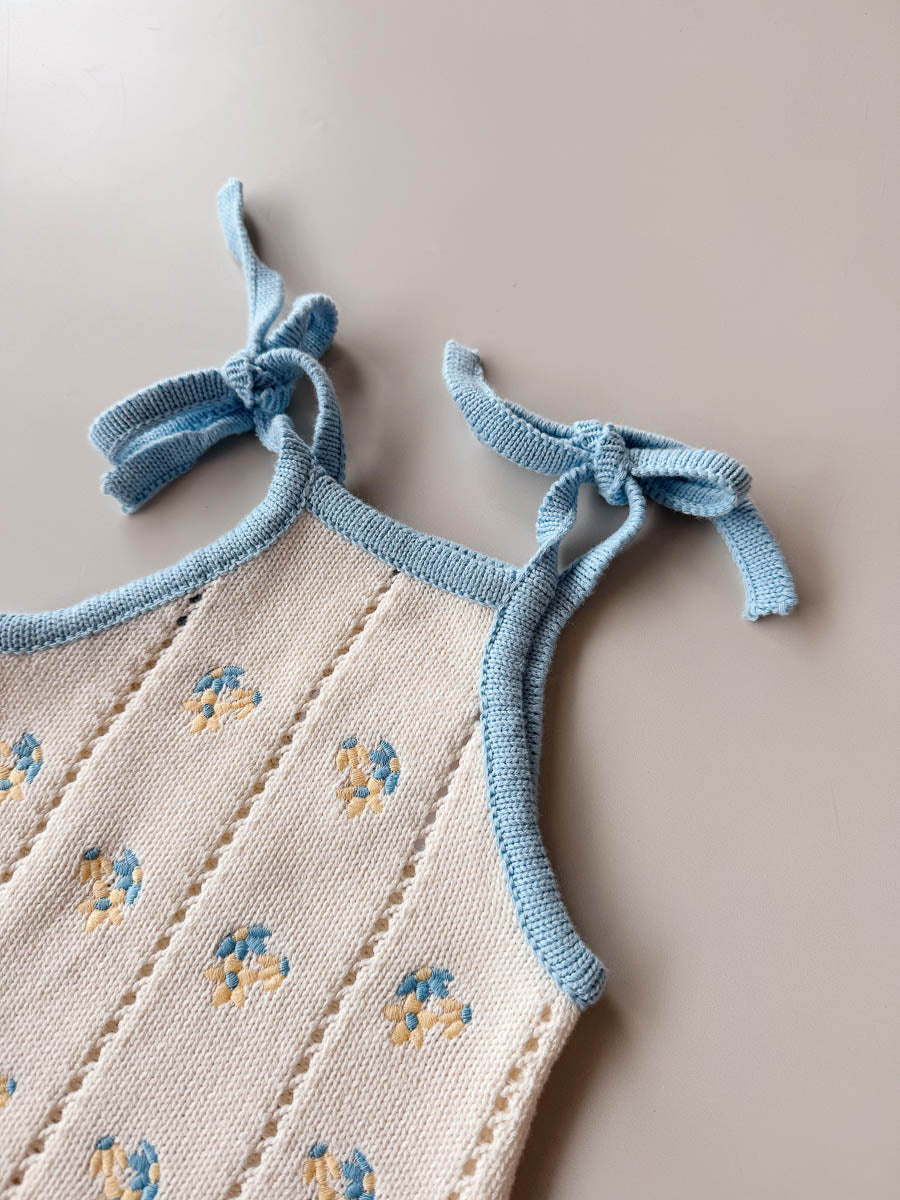 Baby outfit with floral embroidery and blue straps on a beige background