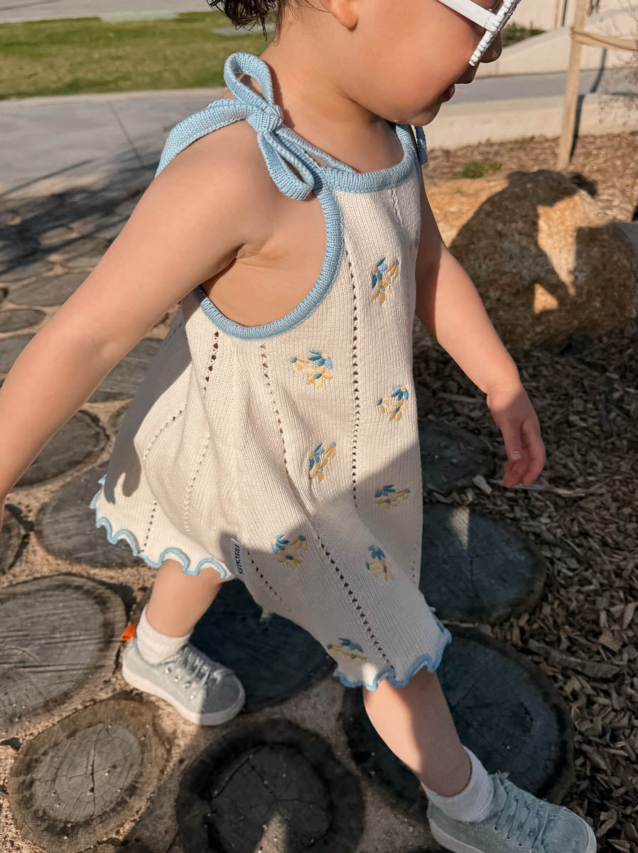 Child wearing a cream dress with light blue and beige floral Embroidery on a stone path.